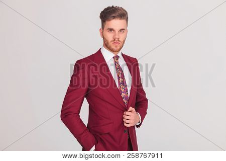 confident elegant young businessman standing on grey background with hand in pocket and the other one holding his coat's button