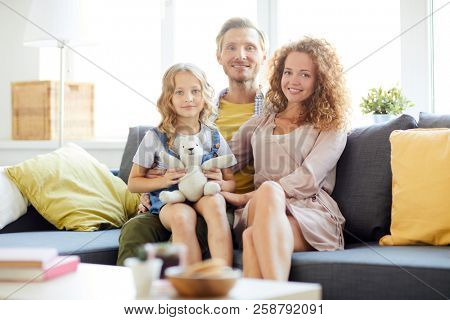 Cute girl with teddybear sitting on her dad knees with mom near by while the family relaxing at home