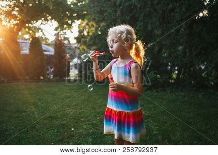 Candid Portrait Of Cute Funny Little Blond Caucasian Child Girl Blowing Soap Bubbles On Home Backyar