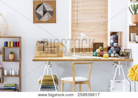 Wooden Chair At Desk With Lamp And Colorful Yarns In Home Office Interior With Poster. Real Photo