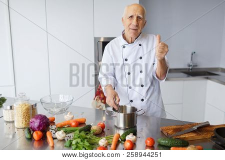 The Aged Chef Man Cooking A Soup And Showing Thumb Up In The Kitchen. The Concepy Of Cooking