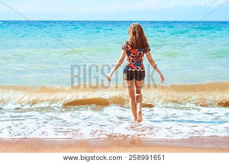 A Young Girl In Shorts Stands With Her Back Against The Background Of Big Waves
