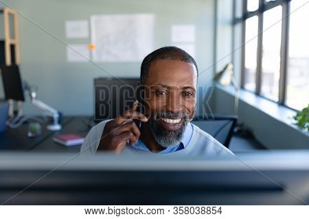 Front view of an African American businessman working in the modern office, sitting by a desk, smiling, talking on his smartphone and using a desktop computer. Social distancing and self isolation in