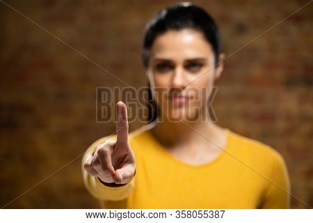 Front view of a Caucasian woman with long dark hair wearing a yellow top with arm raised and hand out in front of her, with her index finger facing camera, touching a virtual interactive screen.