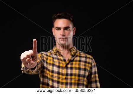 Front view of a Caucasian man with short hair wearing a checked shirt with arm raised and hand out in front of him, with his index finger facing camera, touching a virtual interactive screen on black