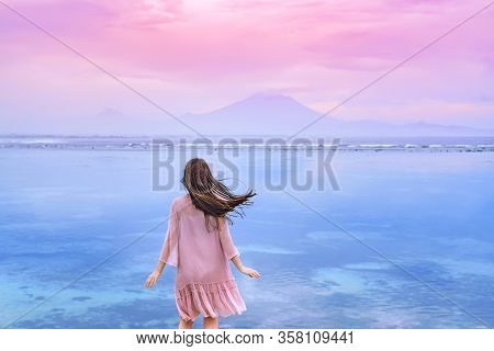 A Young Brunette Woman In A Pink Dress Blown By The Wind Looks At The Ocean And Mountains In The Dis