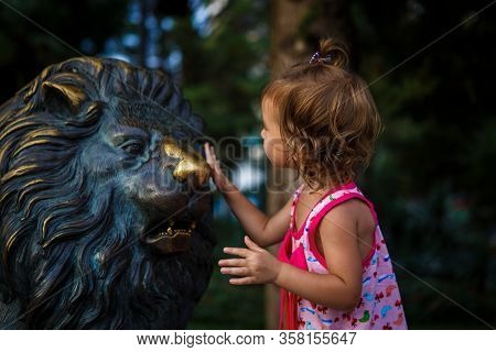 A Little Tourist Girl In A Pink Dress Rubs The Nose Of A Bronze Lion Sculpture. Shiny Nose Of A Bron