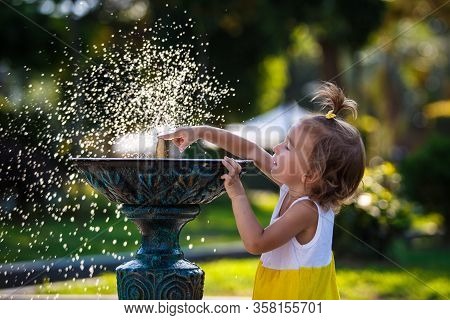 A Little Girl Of 3 Years Old Plays On The Street With A Stream Of Drinking Fountain And Enjoys The S