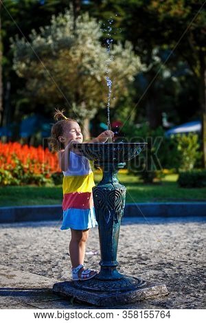 A Little Girl Of 3 Years Old Plays On The Street With A Stream Of Drinking Fountain And Enjoys The S