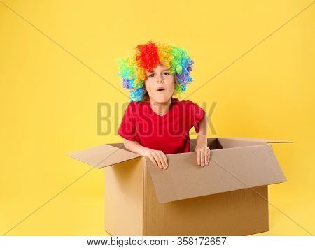 Funny Little Boy In Clown Wig Getting Out Of Cardboard Box On Yellow Background. April Fool's Day