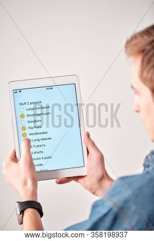 Over shoulder view of young man using packing list in tablet note while putting checkmark in packed stuff