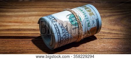 A Roll Of Dollars On A Wooden Textured Table In The Dark Illuminated By A Ray Of Light.