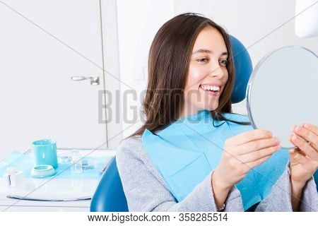 Young Woman Is Sitting In Dental Blue Chair In Clinic, Office. Girl Is Looking Into Mirror On Her Sm