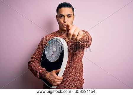 Young brazilian man doing diet to lose weigth holding scale over isolated pink background pointing with finger to the camera and to you, hand sign, positive and confident gesture from the front