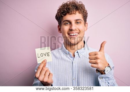 Young blond romantic man with curly hair holdiing paper with love message for valentines day happy with big smile doing ok sign, thumb up with fingers, excellent sign