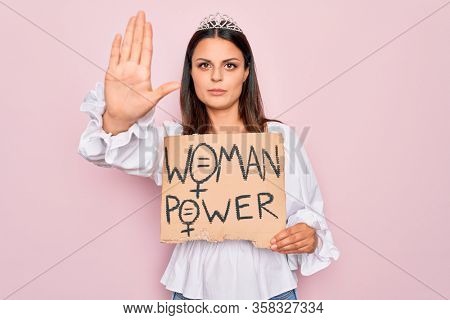 Girl wearing princess tiara asking for women rights holding banner with woman power message with open hand doing stop sign with serious and confident expression, defense gesture