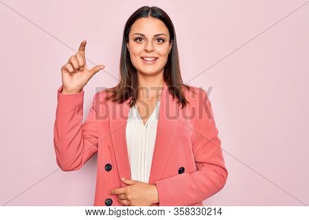 Young beautiful brunette businesswoman wearing elegant jacket over isolated pink background smiling and confident gesturing with hand doing small size sign with fingers looking and the camera. Measure