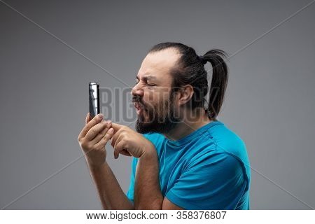 Bearded Man In Blue T-shirt, With Black Hair Combed In Ponytail, Grimacing And Squinting While Using