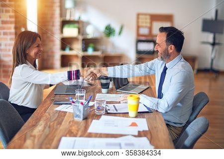 Two middle age business workers smiling happy and confident. Working together with smile on face using laptop doing handsake at the office