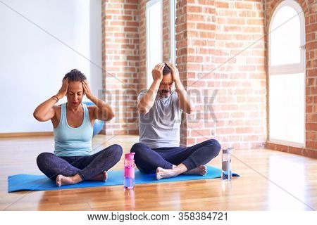 Middle age sporty couple sitting on mat doing stretching yoga exercise at gym suffering from headache desperate and stressed because pain and migraine. Hands on head.