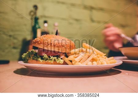 Tasty Delicious Burger On Plate In Cafe. French Fries Beside With Juicy Hamburger. Human Sit On Tabl