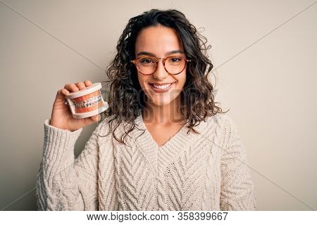 Young beautiful woman with curly hair holding plastic teeth with dental baces with a happy face standing and smiling with a confident smile showing teeth