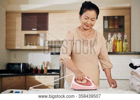Smiling Senior Asian Woman Ironing Clothes At Home During Pandemic Period When She Needs To Stay At 