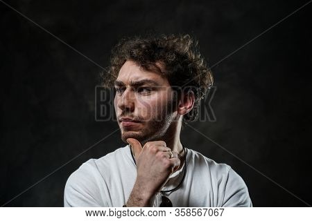 Curly And Hansome Caucasian Man Standing In A Dark Studio On A Grey Background, Wearing Casual White