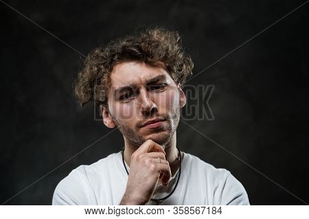 Curly And Hansome Caucasian Man Standing In A Dark Studio On A Grey Background, Wearing Casual White