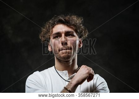 Curly And Hansome Caucasian Man Standing In A Dark Studio On A Grey Background, Wearing Casual White
