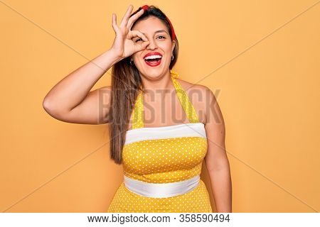 Young hispanic pin up woman wearing fashion sexy 50s style over yellow background doing ok gesture with hand smiling, eye looking through fingers with happy face.