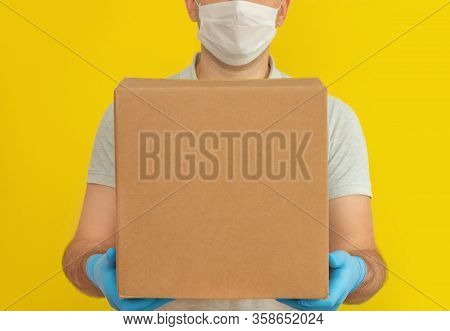 Delivery Man Holding Cardboard Boxes In Medical Rubber Gloves And Mask On Yellow Background. Copy Sp