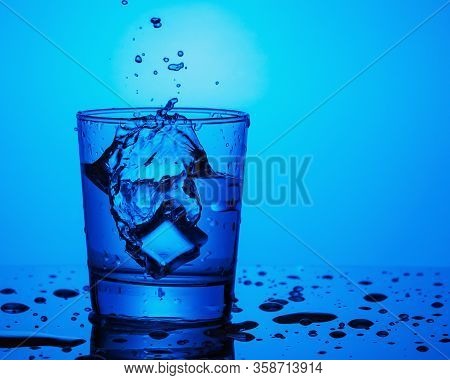 One Glass Of Water With Splashes From Falling Ice Cube On Blue Background