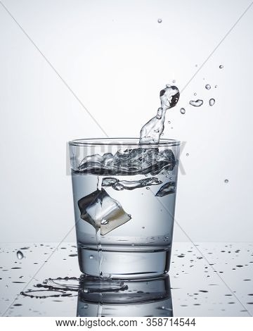 One Glass Of Water With Splashes From Falling Ice Cube On Blue Background