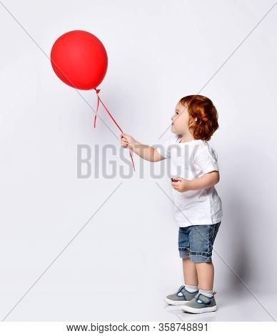 Ginger Toddler Boy Or Girl In White T-shirt, Shoes And Denim Shorts. Child Smiling, Holding Red Ball