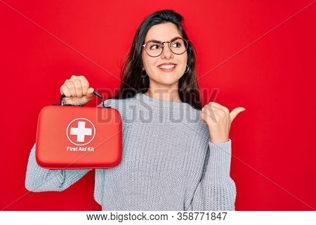Young beautiful girl holding first aid kit medical box over red background pointing and showing with thumb up to the side with happy face smiling