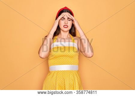 Young beautiful pin up woman wearing 50s fashion vintage dress over yellow background with hand on head for pain in head because stress. Suffering migraine.