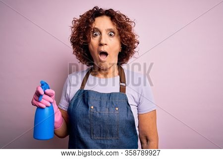 Middle age curly hair woman cleaning doing housework wearing apron and gloves using spayer scared in shock with a surprise face, afraid and excited with fear expression
