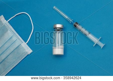 The Syringe Is Next To A Glass Bottle Of Medicine And A Disposable Medical Mask On A Blue Background