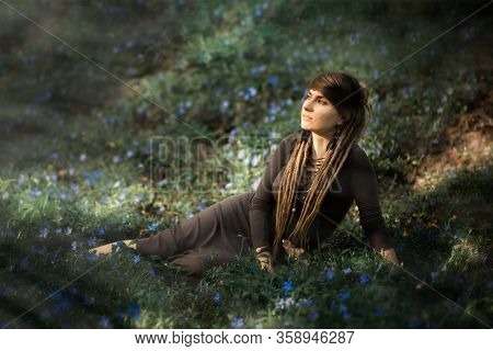 Portrait Of Beautiful Young Woman In Dark Dress With Dreadlocks Sitting On Meadow With Violets Flowe