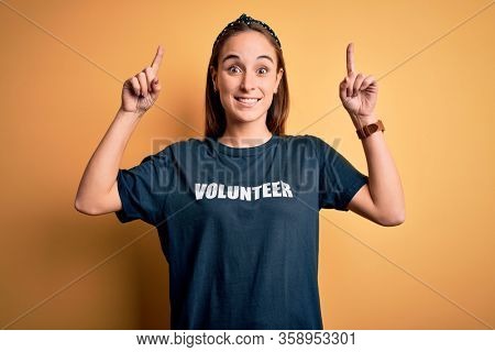 Young beautiful woman wearing volunteer t-shirt doing volunteering over yellow background smiling amazed and surprised and pointing up with fingers and raised arms.