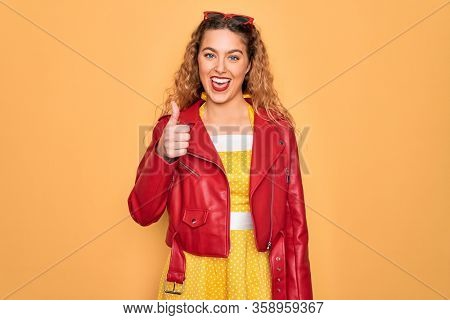 Young beautiful blonde woman pin-up with blue eyes wearing red sunglasses and jacket doing happy thumbs up gesture with hand. Approving expression looking at the camera showing success.