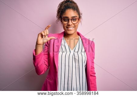 Beautiful african american businesswoman wearing jacket and glasses over pink background smiling and confident gesturing with hand doing small size sign with fingers looking and the camera. Measure