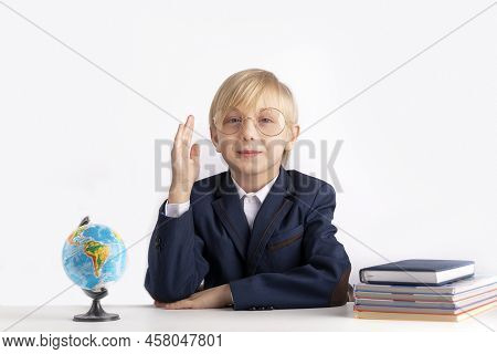 Schoolboy In Large Glasses Sits At Table With Textbooks With His Hand Raised. Excellent Student. Boy