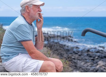 Caucasian Bearded Senior Man With Hat Sitting At The Beach Looking At The Horizon While Smoking A Ci