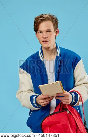 A Pensive Student In A Blue Stylish Bomber Jacket Stands With A Notebook In His Hands And Looks Away
