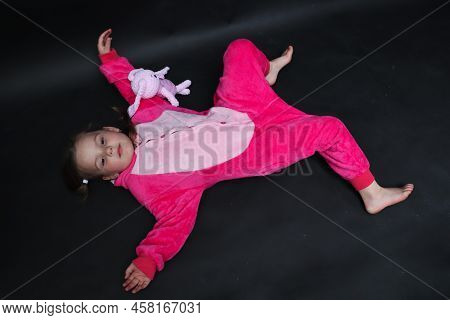 Overindulgence. The Baby Is Playing. The Child Lies On A Black Background In An Embrace With A Hare.