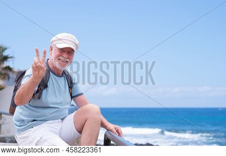 Attractive Cheerful Elderly Bearded Man With Hat And Backpack Sitting Near The Beach By The Sea Look