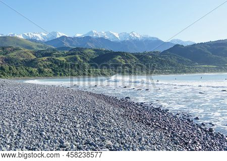 Stony Mangamaunu, Beach Near Kaikoura With Surf Rolling In And Surfers Under Backdrop Of Coastal Ran