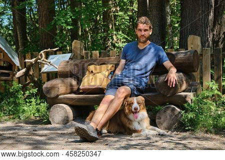 Kivach Nature Reserve, Karelia. Man Is Sitting In Park On Wooden Bench Made Of Logs And Dog Is Lying